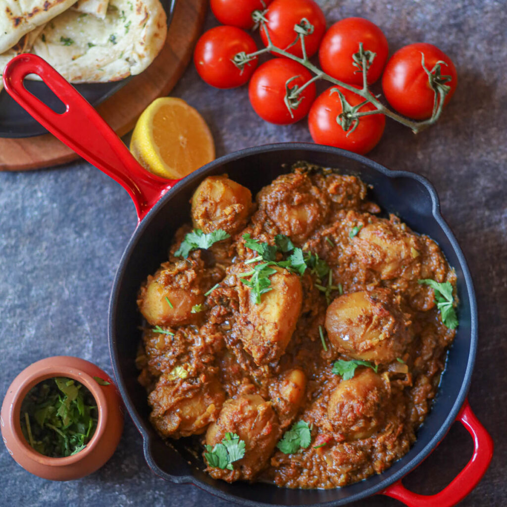 dum aloo on red skillet with tomatoes on the side